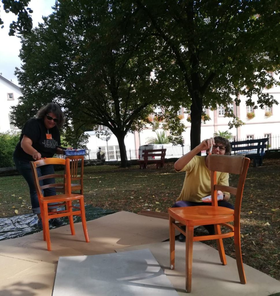 Zur Vorbereitung einer öffentlichkeitswirksamen Aktion bekommen gebrauchte Stühle einen orangefarbenen Anstrich verpasst.